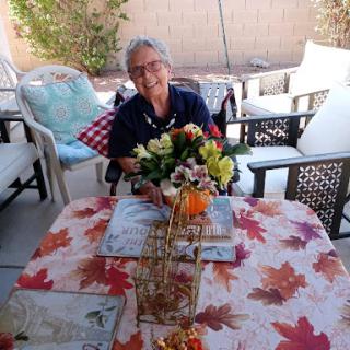 An elderly woman enjoying a sunny day outdoors, surrounded by autumn decorations and a beautiful floral arrangement.
