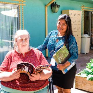A caregiver interacting with an elderly woman in a vibrant outdoor setting, as they share a moment over a book.