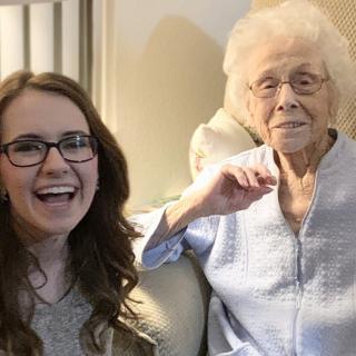 A young woman with glasses smiles next to an elderly woman sitting indoors.