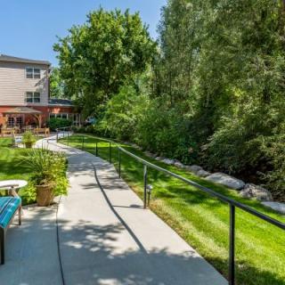 A scenic outdoor view with a garden path winding past lush greenery and trees, adjacent to a building on a sunny day.