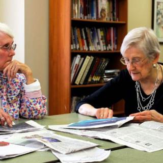 Two elderly women reading newspapers and discussing in a library.