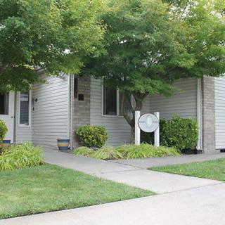 A residential building with a landscaped front yard, featuring trees and bushes along a concrete sidewalk.