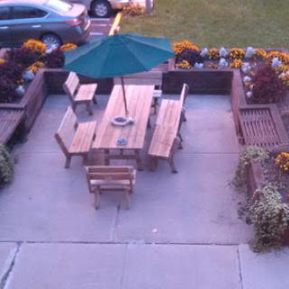 A cozy outdoor seating area with a wooden table, chairs, and a green umbrella surrounded by flowers.