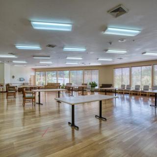 An empty community center room with tables and chairs arranged on a wooden floor, illuminated by fluorescent lights.