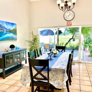 A bright dining room featuring a long table with a floral tablecloth, surrounded by chairs. The room showcases artwork on the wall, a decorative chandelier, and a clock above the sliding glass door that opens to a garden view.