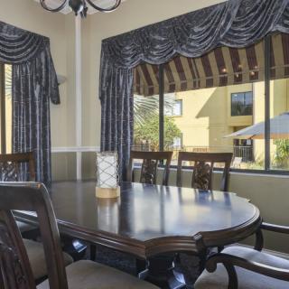 A dining room featuring a large wooden table and upholstered chairs, with windows adorned by dark curtains.