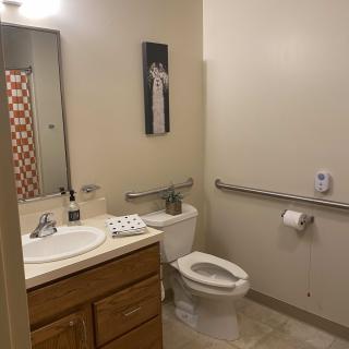 A clean bathroom with a sink, mirror, toilet, and safety handrail.
