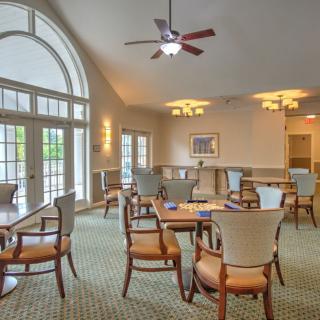 A well-lit dining area with tables and chairs positioned under a ceiling fan and large windows.