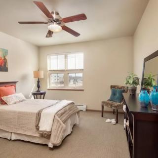 A modern and well-decorated bedroom featuring a bed with white linens, a ceiling fan, artwork on the wall, and natural light streaming through the windows.