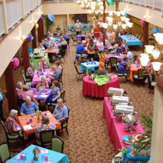 An indoor celebration with multiple tables, decorated with colorful tablecloths and balloons, where people are enjoying a buffet.