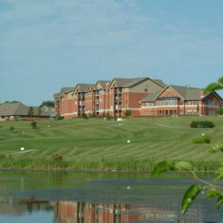 A large building sits by a calm lake, surrounded by green grass and under a blue sky.