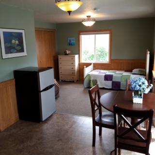 A cozy bedroom with a small refrigerator, bed, table, and dresser next to a window.