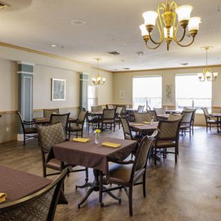 An empty dining room with neatly arranged tables and chairs, illuminated by chandeliers and natural light from large windows.