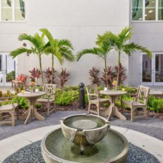 A serene courtyard featuring a central fountain, surrounded by tables and chairs, with palm trees and plants enhancing the tranquil atmosphere.