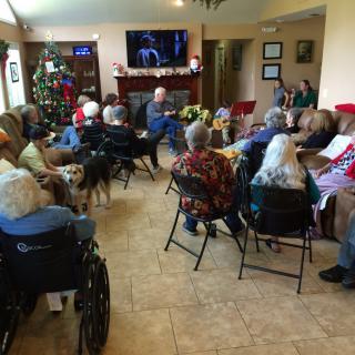 A group of elderly individuals gathered in a common room, watching television with festive holiday decorations around.