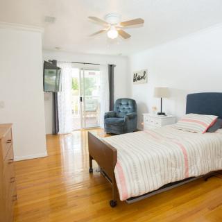 A bright bedroom with a bed, armchair, and a ceiling fan, featuring hardwood floors and sliding glass doors.