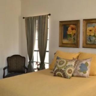 A cozy bedroom with a yellow bedspread and decorative pillows, featuring artwork on the wall and curtains by the window.