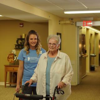 A caregiver assists an elderly woman using a walker in a nursing home.