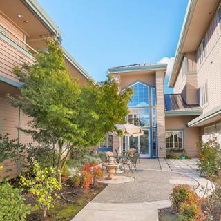 A view of a modern building's courtyard featuring trees, plants, and outdoor furniture under a clear blue sky.