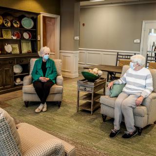 Two seniors wearing masks sit and converse in a cozy living room setting.
