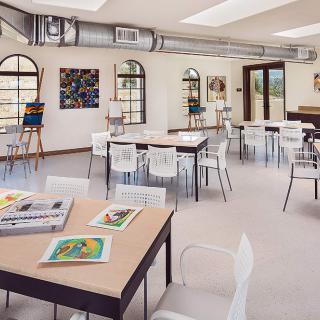 A bright and spacious art classroom with tables, chairs, and easels, featuring windows that allow natural light to fill the room.