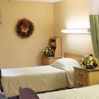 A simple hospital room featuring two twin beds, adorned with floral arrangements and a wall-mounted wreath.