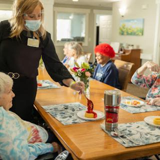 A caregiver serves a meal to elderly residents in a warm and inviting dining setting.