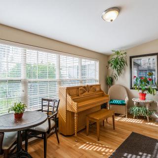 A cozy living room filled with natural light, featuring a piano and a small dining set surrounded by lush plants.