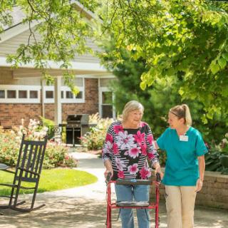 A caregiver assists an elderly woman with a walker in a sunny garden setting.
