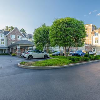 A building with a driveway and parked cars surrounded by trees on a sunny day.
