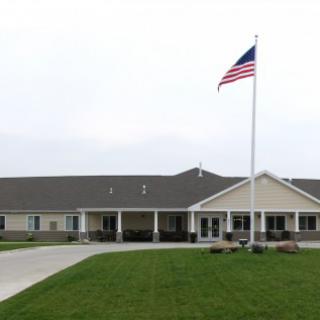 A building with an American flag on a tall pole, surrounded by a green lawn and driveway under an overcast sky.