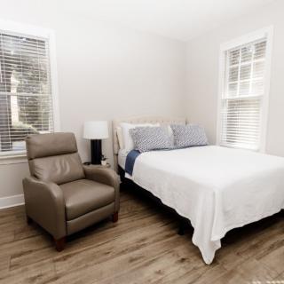 A minimalist bedroom with a bed, armchair, and lamp, featuring large windows and hardwood flooring.