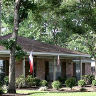 A suburban brick house surrounded by trees and a well-maintained garden, with flags displayed at the front.