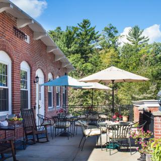 A sunny outdoor patio with tables, chairs, and umbrellas next to a brick building surrounded by trees.