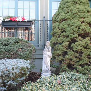 A garden scene featuring a statue surrounded by shrubbery, with a large window and a planter box filled with flowers in the background.