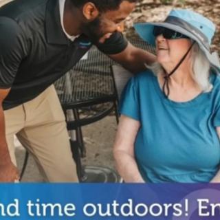 A senior woman in a sun hat and sunglasses enjoys a conversation outdoors with a man.