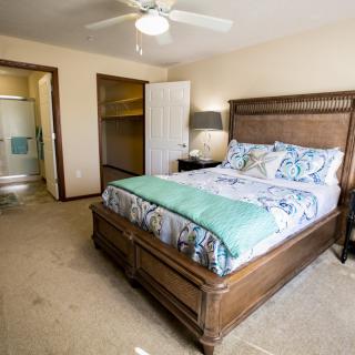 A cozy bedroom with a wooden bedframe, colorful bedding, a nightstand with a lamp, and an open closet door, featuring a ceiling fan for comfort.