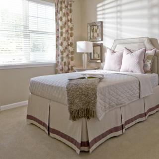 A cozy bedroom featuring a neatly made bed with decorative pillows, a bedside lamp, and a window with light filtering through sheer curtains.