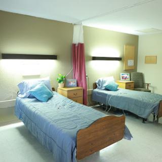 A view of a patient room featuring two hospital beds with bedside tables and soft lighting.