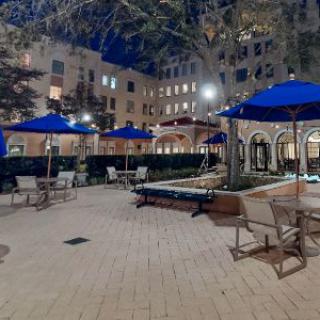 A nighttime view of a well-lit outdoor patio area with tables and chairs under blue umbrellas, surrounded by buildings.