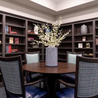 A cozy library corner with a round table and chairs, surrounded by bookshelves, featuring a decorative vase with flowers.