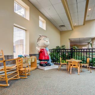 A children's play area featuring a large character statue, small tables and chairs, wooden rocking chairs, and windows allowing natural light.