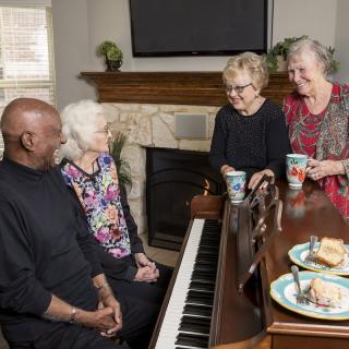 A group of seniors enjoying coffee and conversation around a piano.