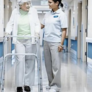 A nurse assists an elderly woman using a walker in a hospital corridor.