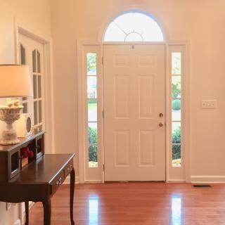 A well-lit foyer with a white front door, wooden flooring, and a side table with a lamp.