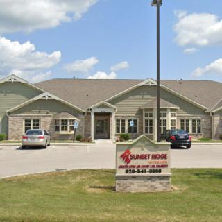 A commercial building with several cars parked in front and a sign displayed on the lawn under a partly cloudy sky.