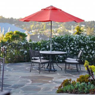 A peaceful outdoor patio with a table and chairs under a red umbrella, surrounded by lush greenery.