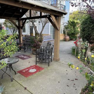 A cozy outdoor patio area with chairs and tables under a wooden canopy, surrounded by lush garden plants.