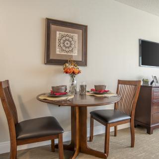 A cozy dining area with a round table set for two, featuring elegant chairs and a decorative vase of flowers, enhanced by wall art and a flat-screen TV.