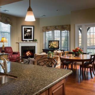 A spacious and elegantly designed living room featuring an open floor plan with a kitchen island and dining area, complemented by natural light from large windows and cozy setup around the fireplace.